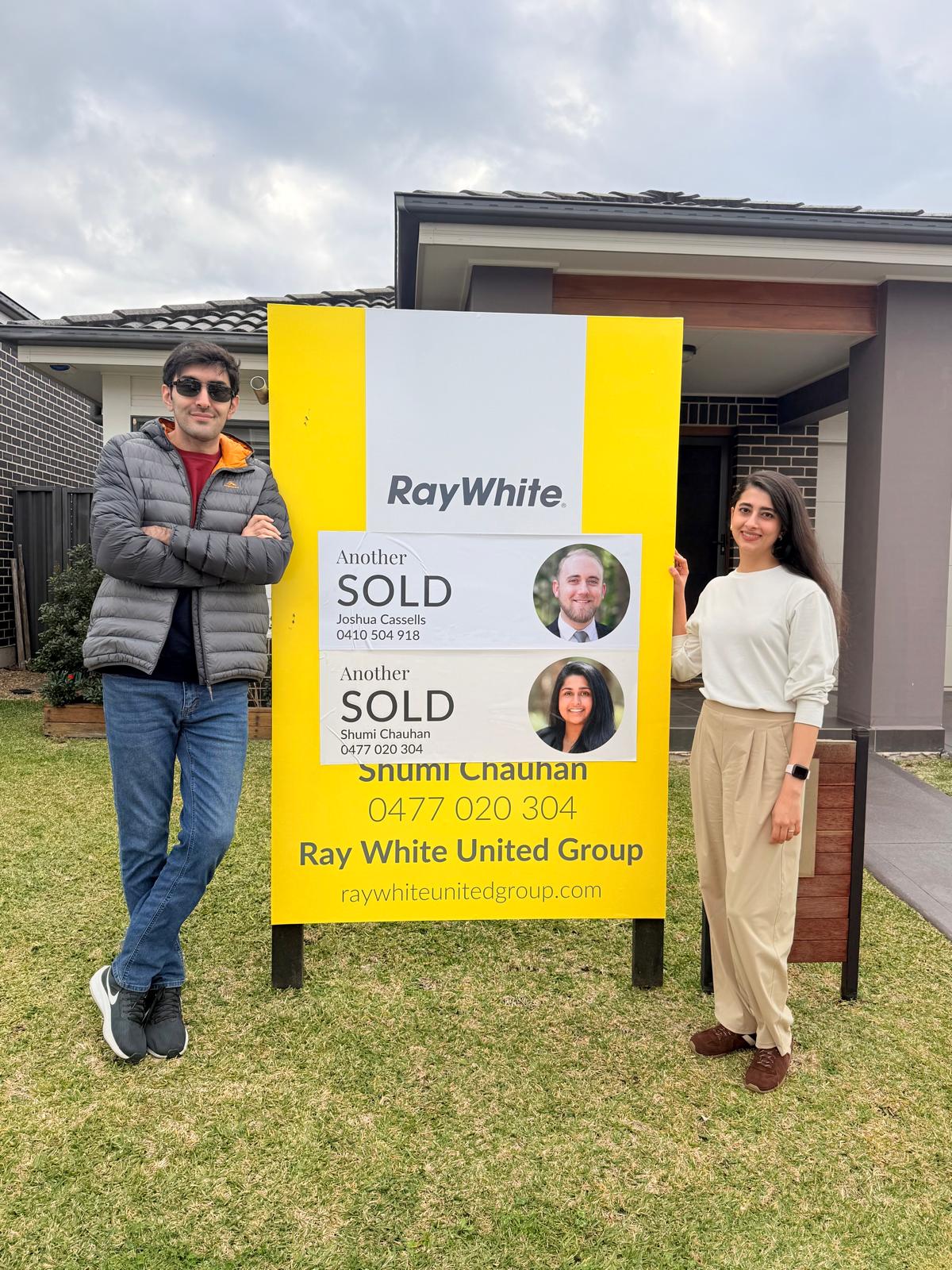 Two sellers beside a Sold sign at their home
