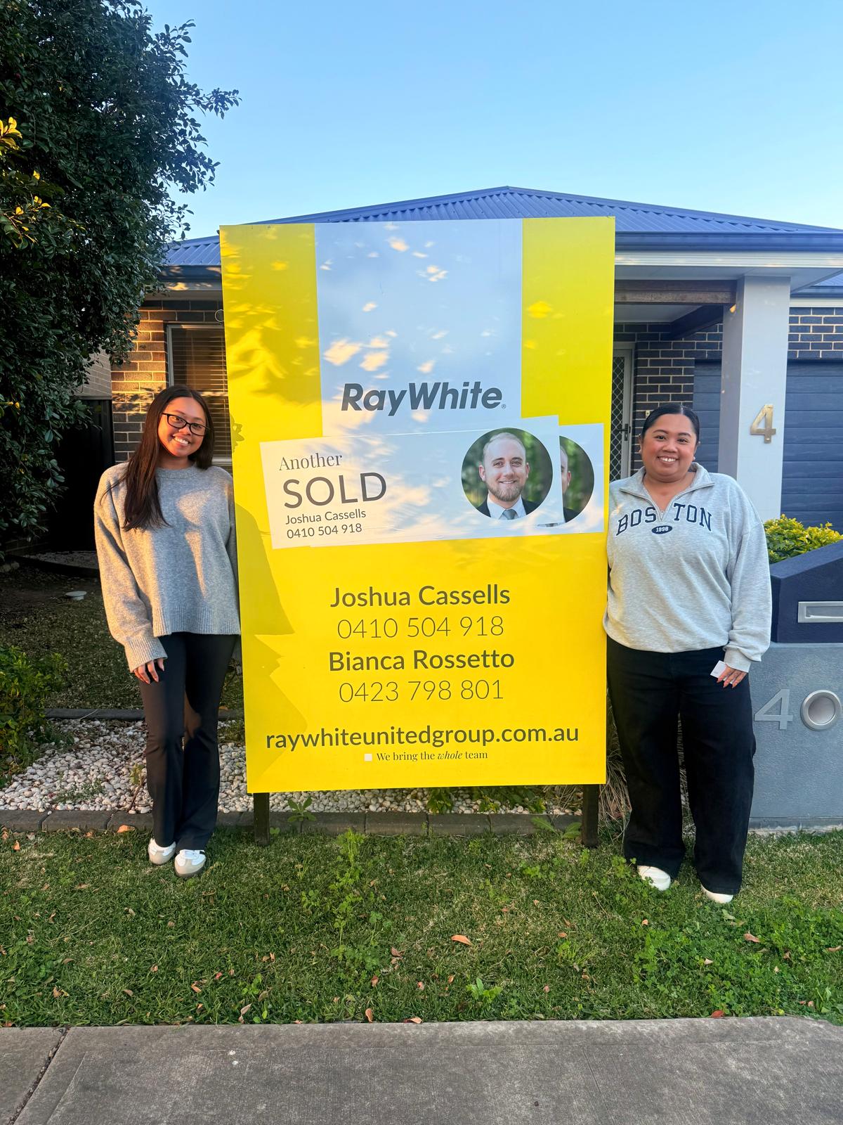 Two sellers beside a Sold sign at their home in the evening