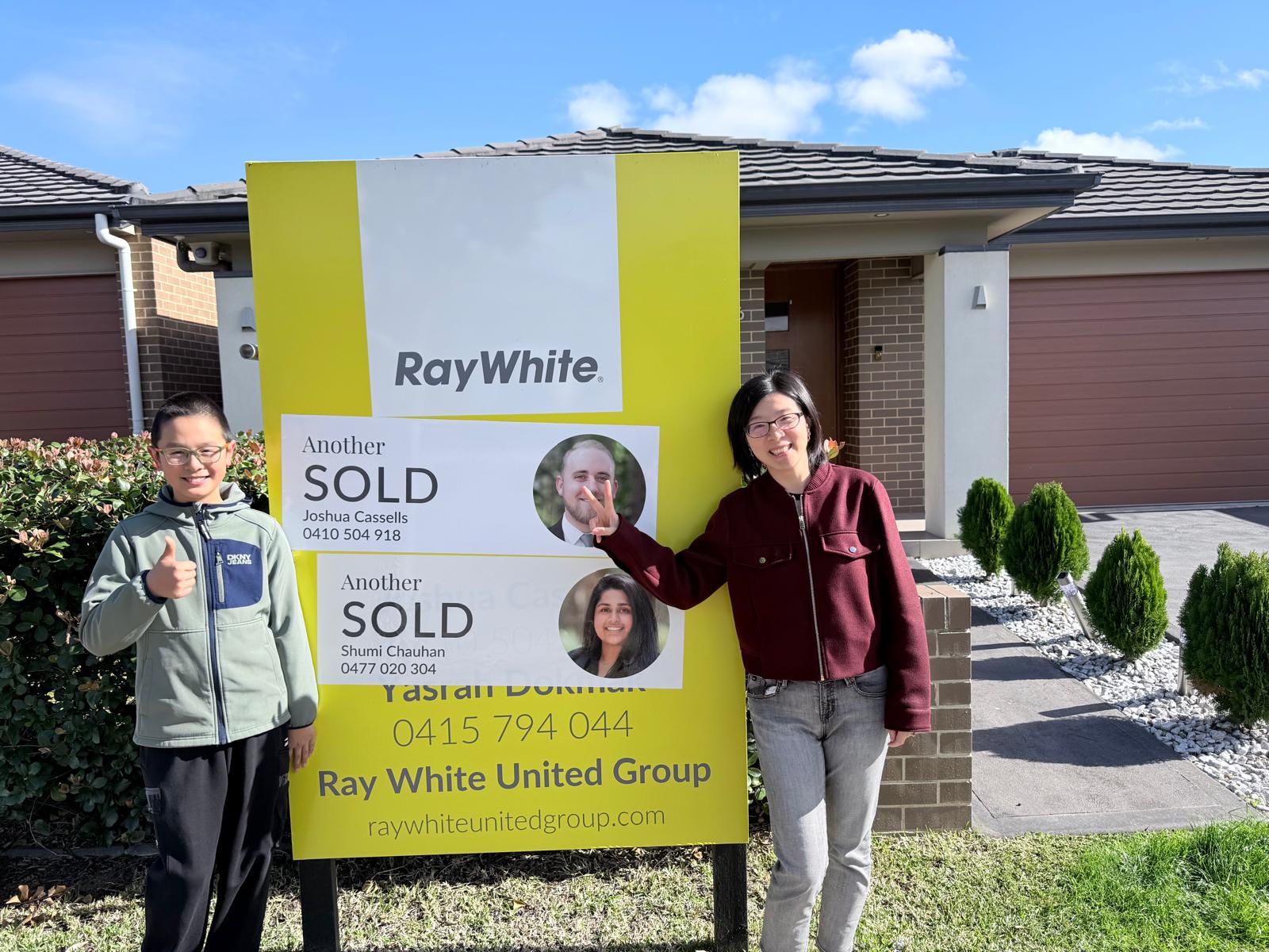 A seller and her son beside a Sold sign at their home